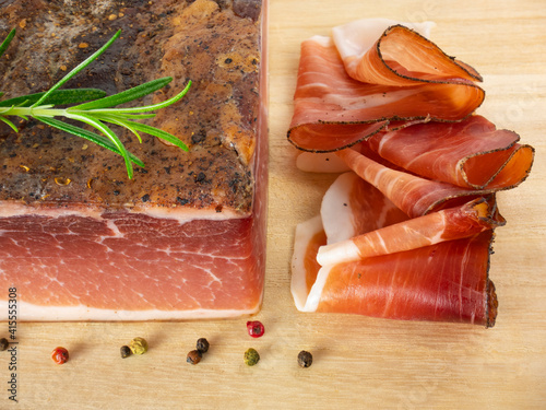 Speck with slices on the cutting board. Typical South Tyrolean smoked bacon. Sliced raw ham. Dry cured meat. Traditional cold cuts, Italian speck with rosemary and pepper. Flat lay.