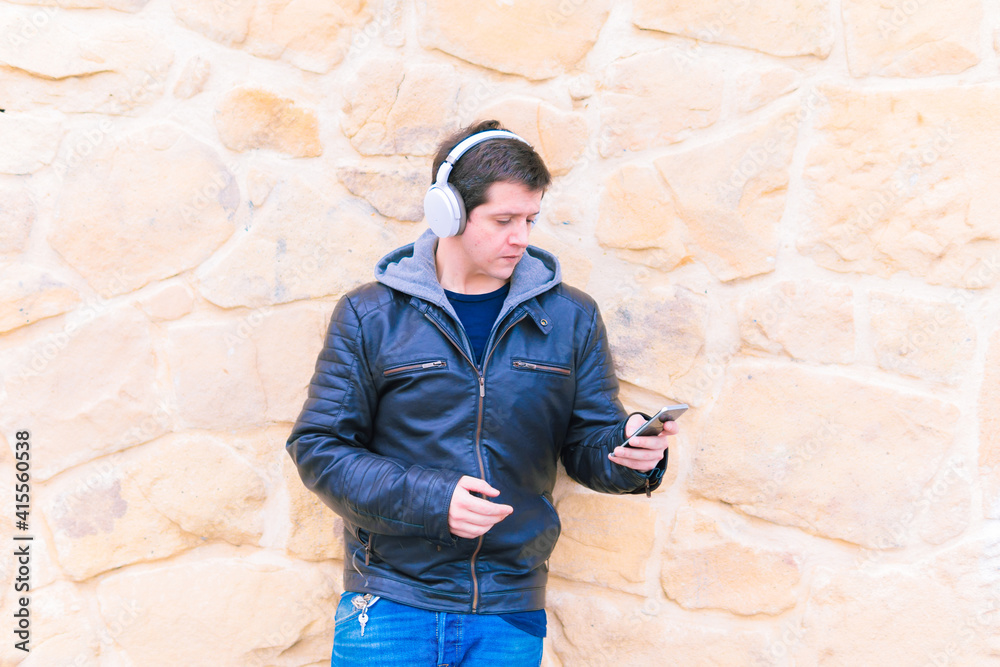 Attractive young middle-aged man leaning on stone wall wearing wireless music headphones and leather jacket looking at smartphone