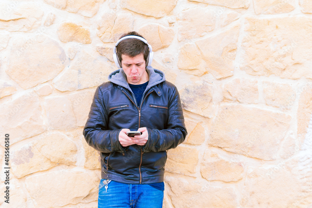 Attractive young middle-aged man leaning on stone wall wearing wireless music headphones and leather jacket looking at smartphone
