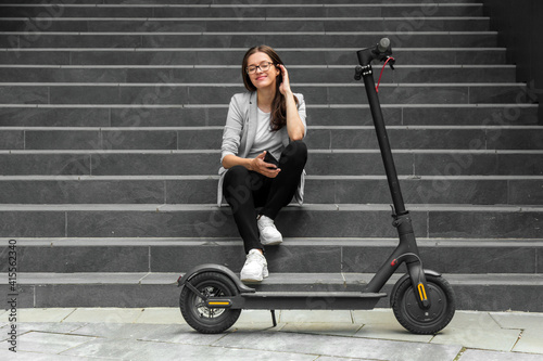 Young woman with smartphone is sitting on the stairs near electric scooter 