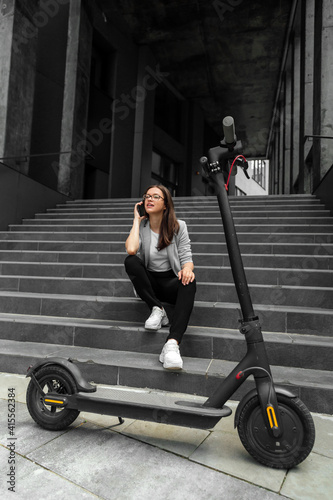 Young woman sitting on stairs talking on smartphone near electric scooter