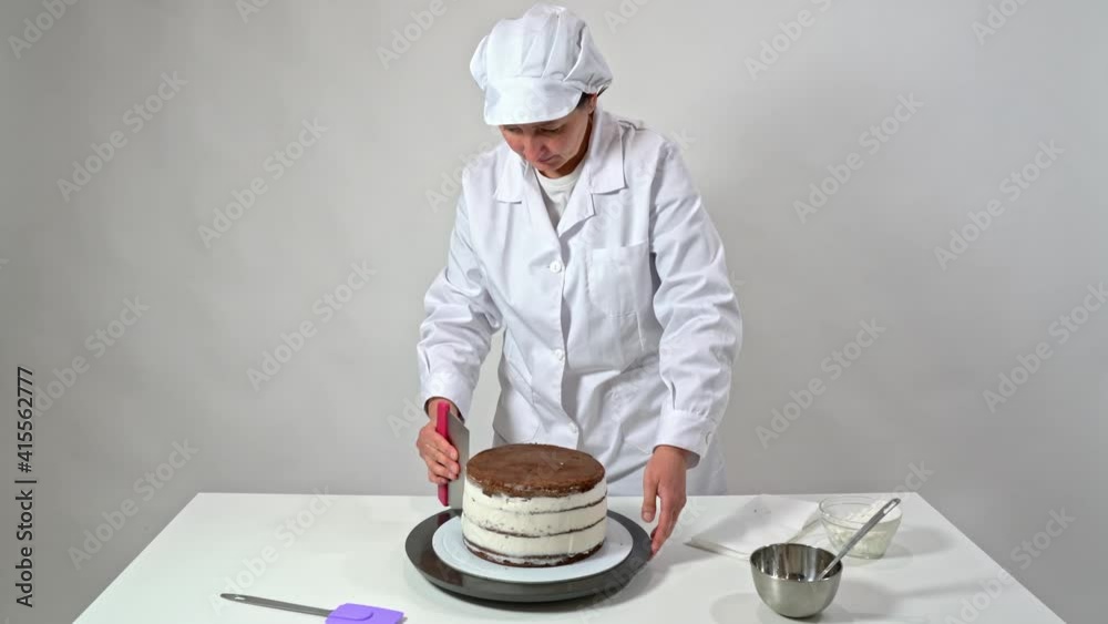 Baker removes the excess cream from the sides of a chocolate cake by rotating the stand and using a spatula. Baker removes the excess cream from the sides of a chocolate cake by rotating the stand and using a spatula.