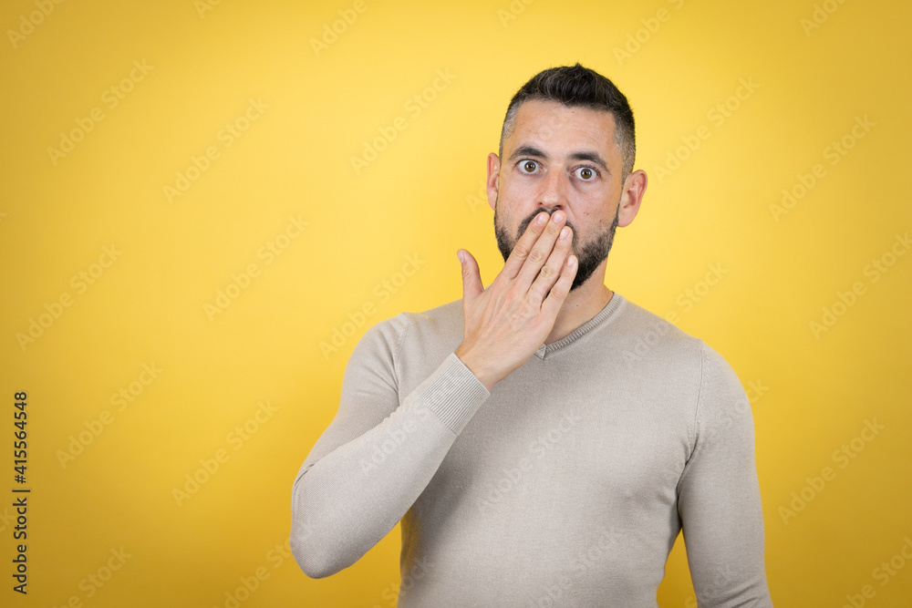 Handsome man with beard wearing sweater over yellow background covering mouth with hand, shocked and afraid for mistake. surprised expression Handsome man with beard wearing sweater over yellow background covering mouth with hand, shocked and afraid for mistake. surprised expression