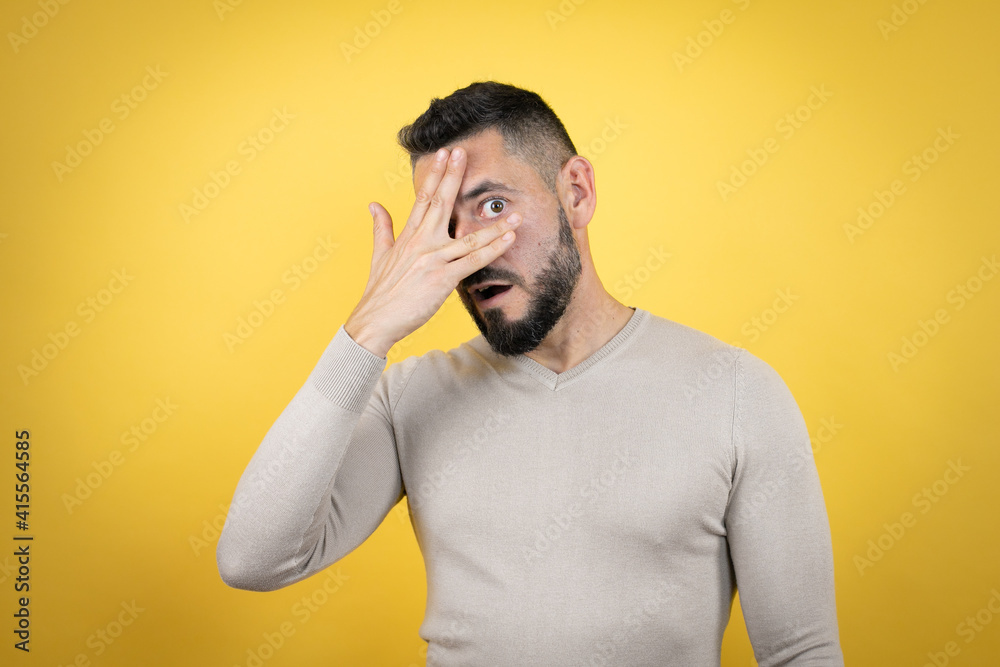 Handsome man with beard wearing sweater over yellow background peeking in shock covering face and eyes with hand, looking through fingers with embarrassed expression Handsome man with beard wearing sweater over yellow background peeking in shock covering face and eyes with hand, looking through fingers with embarrassed expression