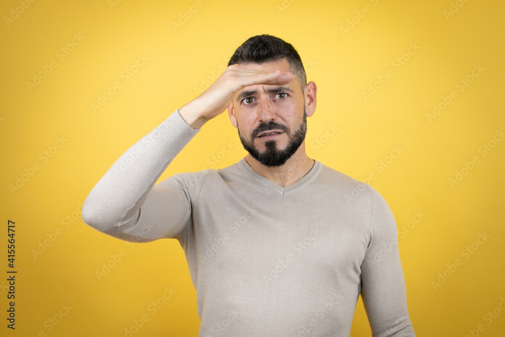 Handsome man with beard wearing sweater over yellow background very happy and smiling looking far away with hand over head. searching concept.