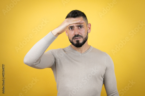 Handsome man with beard wearing sweater over yellow background very happy and smiling looking far away with hand over head. searching concept.