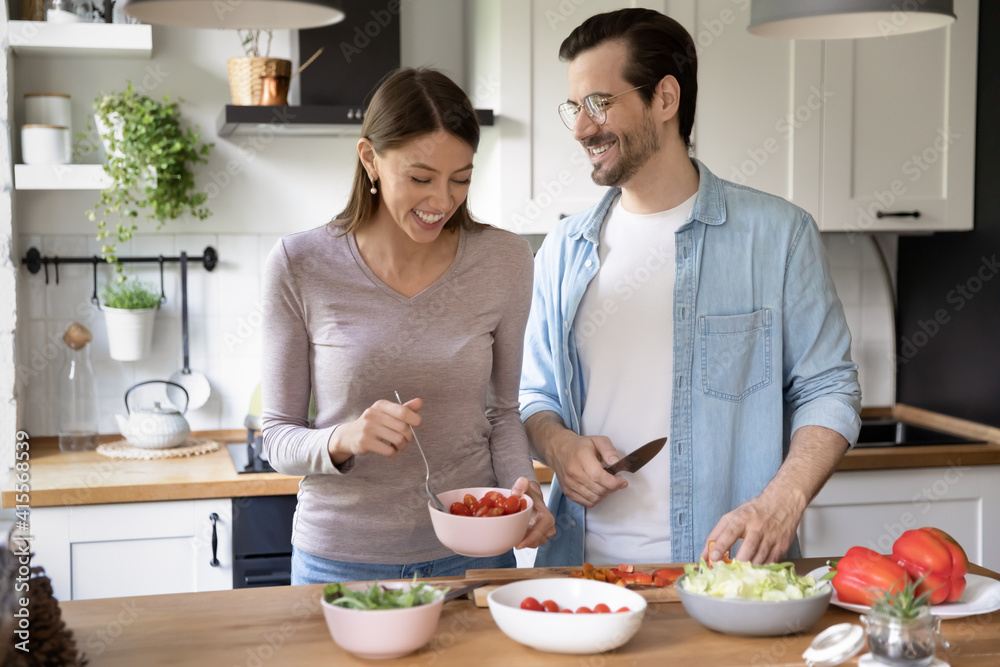 Overjoyed millennial man and woman have fun cooking healthy tasty vegetarian salad in kitchen together. Smiling young Caucasian couple prepare delicious vegetable diet food in own cozy house.