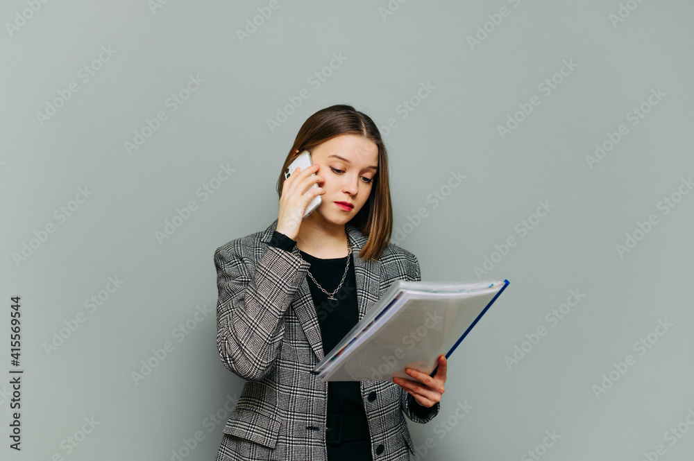 Fototapeta premium Young female office worker in smart casual clothes stands on a gray background and calls on a smartphone, reads documents with a serious face. Lady is in business, isolated.