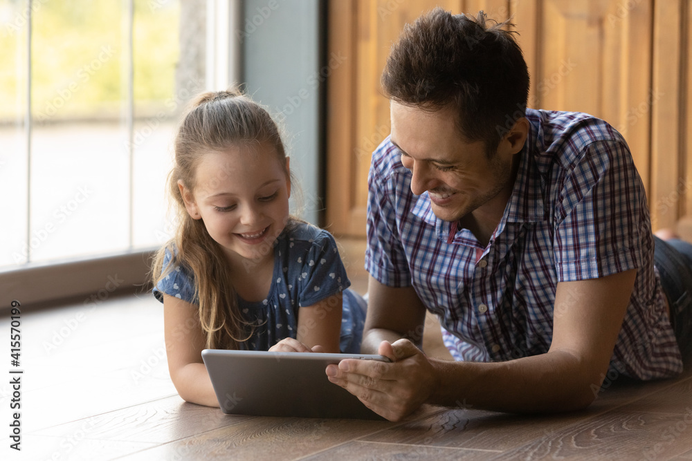 Caring father and little daughter having fun with tablet together, lying on warm floor with underfloor heating, smiling dad with adorable child browsing apps, playing game, watching video at home