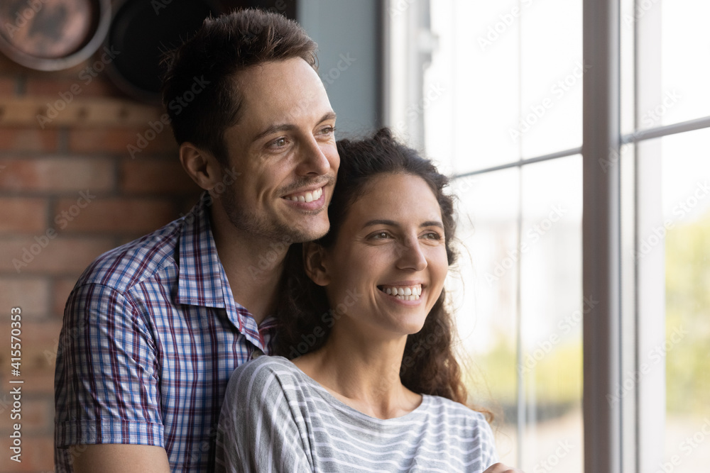 Close up smiling man and woman hugging, looking out window, standing at home, happy dreamy young couple visualizing future together, excited wife and husband planning, dreaming and thinking
