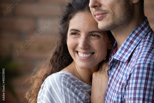 Close up smiling beautiful dreamy woman hugging man, looking to aside, happy young wife visualizing good future with husband, young couple enjoying tender moment, planning, standing at home