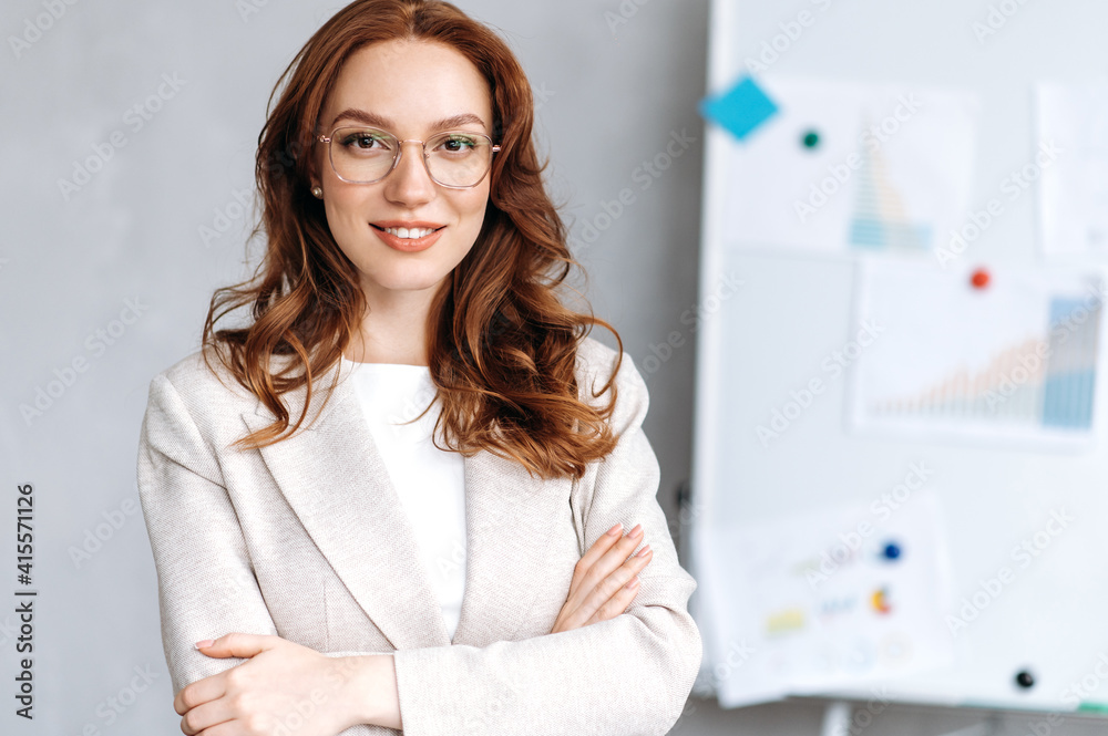 Portrait of a beautiful red-haired young business woman dressed in stylish formal suit and glasses, standing with crossed arms in the office, looking and smiling friendly at the camera Portrait of a beautiful red-haired young business woman dressed in stylish formal suit and glasses, standing with crossed arms in the office, looking and smiling friendly at the camera