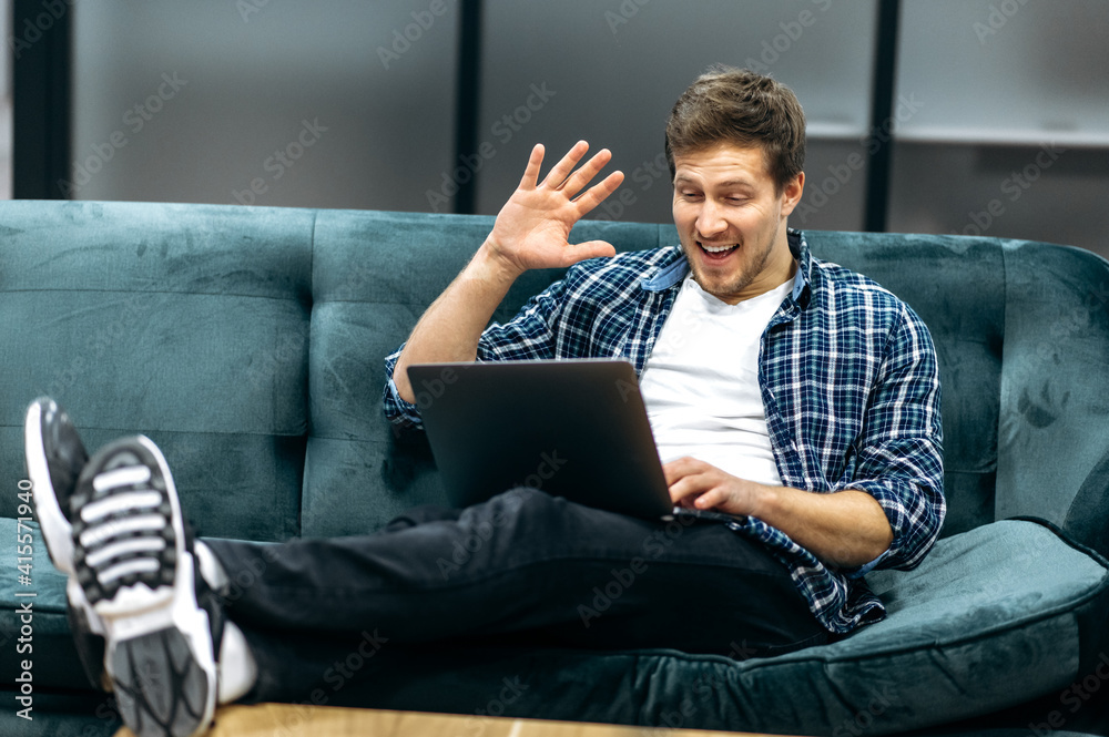 Joyful male freelancer or student in casual stylish wear sitting in modern office, using laptop. The guy communicates by video call with colleagues or friends, smiling, communication concept