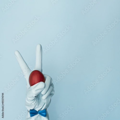 Gloved hand holds an Easter red egg on a pastel, trendy blue background. Dressy rabbit with copy space. Minimal Easter concept. Corona virus style.
