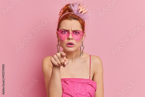 Angry redhead young woman wears stylish sunglasses and dress indicates directly at camera with annoyed face expression sees something stunning isolated over pink background. Monochrome shot. Hey you
