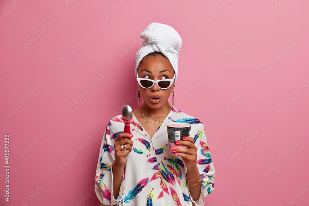 Surprised dark skinned woman looks surprisingly aside wears domestic robe bath towel wrapped on head eats delicious cold ice cream holds spoon poses in sungasses indoor against pink background