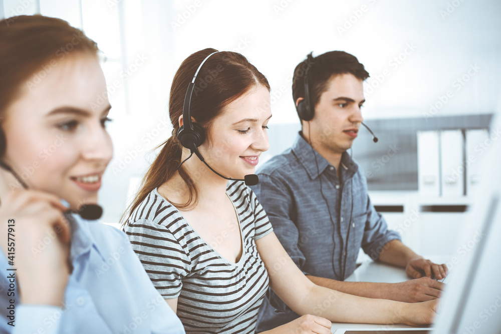 Casual dressed young woman using headset and computer while talking with customers online. Group of operators at work. Call center, business concept Casual dressed young woman using headset and computer while talking with customers online. Group of operators at work. Call center, business concept