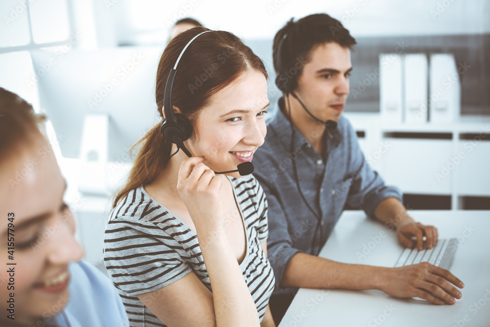 Casual dressed young woman using headset and computer while talking with customers online. Group of operators at work. Call center, business concept