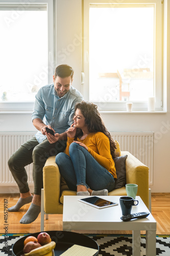Smiling couple talking and looking in mobile phone. Loving couple laughing and watching something on smartphone together at home