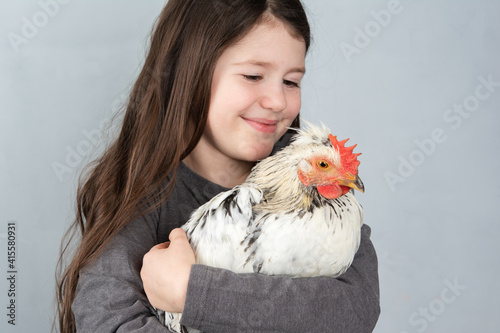 Fotografija Little girl holdinig white cockerel with black feathers, smiling, on grey backgr