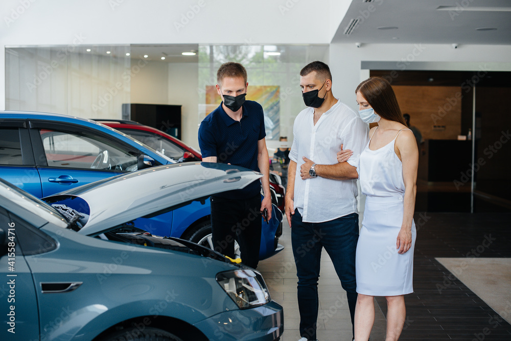 Young couple in masks selects a new vehicle and consult with a representative of the dealership in the period of the pandemic. Car sales, and life during the pandemic Young couple in masks selects a new vehicle and consult with a representative of the dealership in the period of the pandemic. Car sales, and life during the pandemic