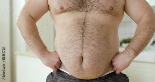 The concept of human obesity. Shirtless overweight man with hairing stomach scratching his tummy, front view. He's trying to flex his abs muscles.
