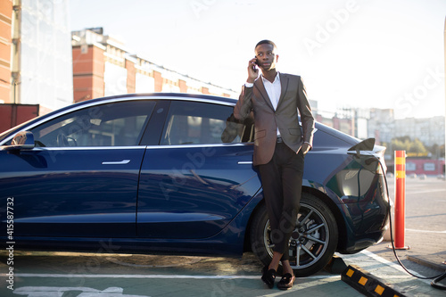 Electric car charging concept. Full length portrait of young handsome dark skinned man, standing at the charging station and talking phone whie waiting for his car charging © sofiko14