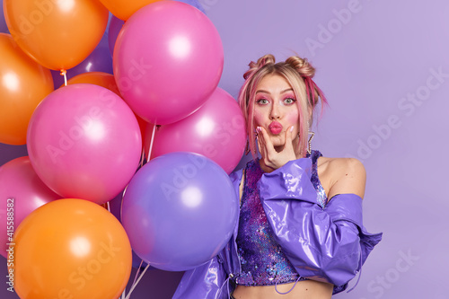 Stylish European woman keeps lips folded dressed in fashionable jacket shows demonstrates bare shoulder holds colorful inflated balloons decorates hall for party isolated over purple background