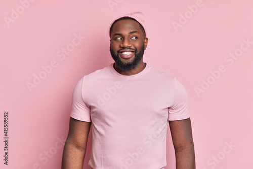 Cheerful bearded black man smiles broady looks curiously aside has white even teeth wears hat and t shirt in one tone with background. Pleased Afro American guy notices something interesting photo