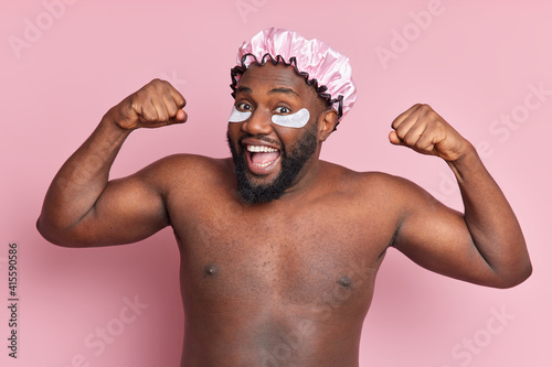 Horizontal shot of positive dark skinned bearded man prepares for taking shower raises arms shows biceps feels amused wears waterproof hat applies collagen pads under eyes stands shirtless indoor