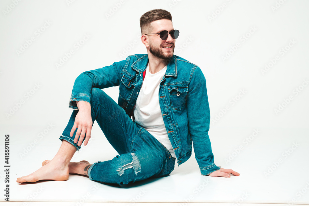 Fototapeta premium Portrait of handsome smiling stylish hipster lambersexual model.Man dressed in jacket and jeans clothes. Fashion male sitting on grey background in studio in sunglasses
