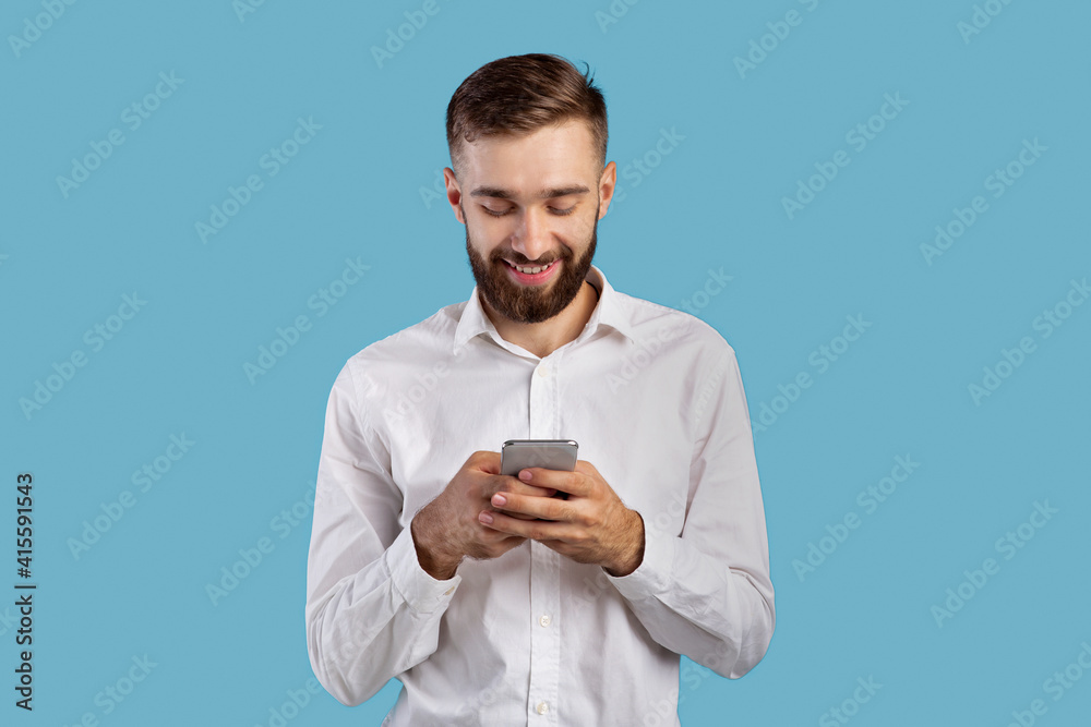 Young man looking at smartphone screen, browsing internet, playing online game, doing shopping on blue background Young man looking at smartphone screen, browsing internet, playing online game, doing shopping on blue background