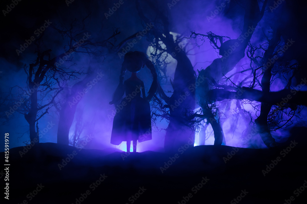 Fototapeta premium Horror Halloween decorated conceptual image. Alone girl with the light in the forest at night. Silhouette of girl standing between trees with surreal light on background. Selective focus.