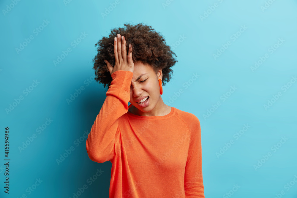 Frustrated dark skinned young woman keeps hand on forehead regrets wrong doing feels stressed dressed in orange sweater isolated over blue background. Upset Afro American girl facepalming indoor