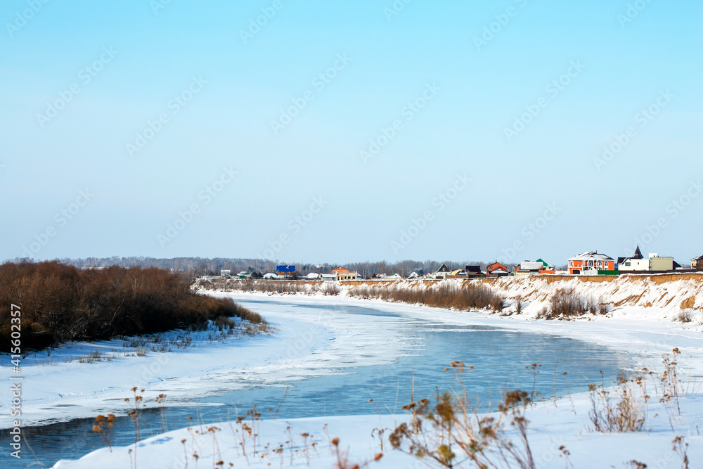
Frosty winter panoramic landscape with forest river on a sunny February morning. Village houses by the river in winter