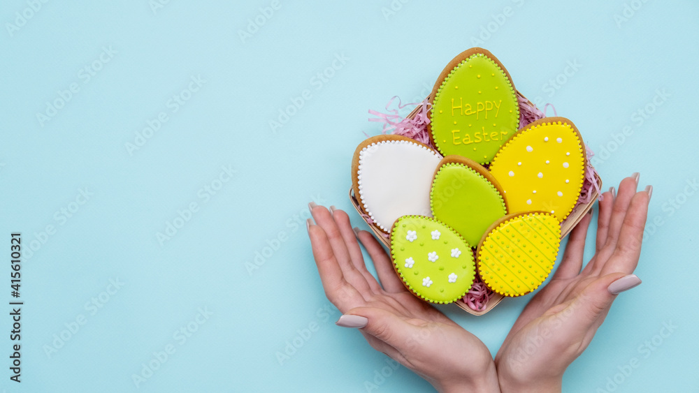 Happy Easter. Handmade sweet gift. Bakery food art. Celebration symbol. Bright green yellow gingerbread cookies eggs in basket in woman open hands isolated on blue empty space background. Happy Easter. Handmade sweet gift. Bakery food art. Celebration symbol. Bright green yellow gingerbread cookies eggs in basket in woman open hands isolated on blue empty space background.