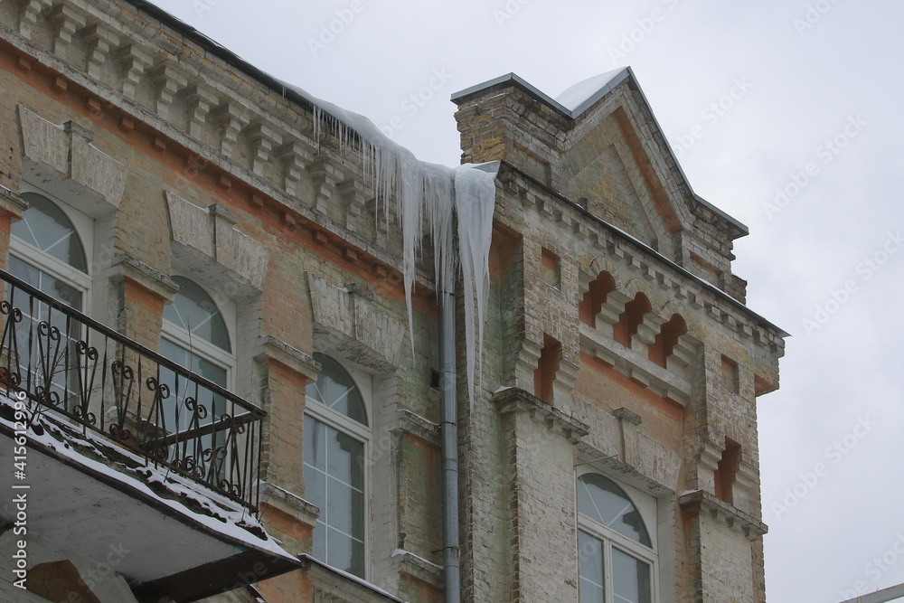 Big, sharp icicles and melted snow hanging from eaves of roof. Dangerous icicles on the roof. Danger of being near a building. City landscape.