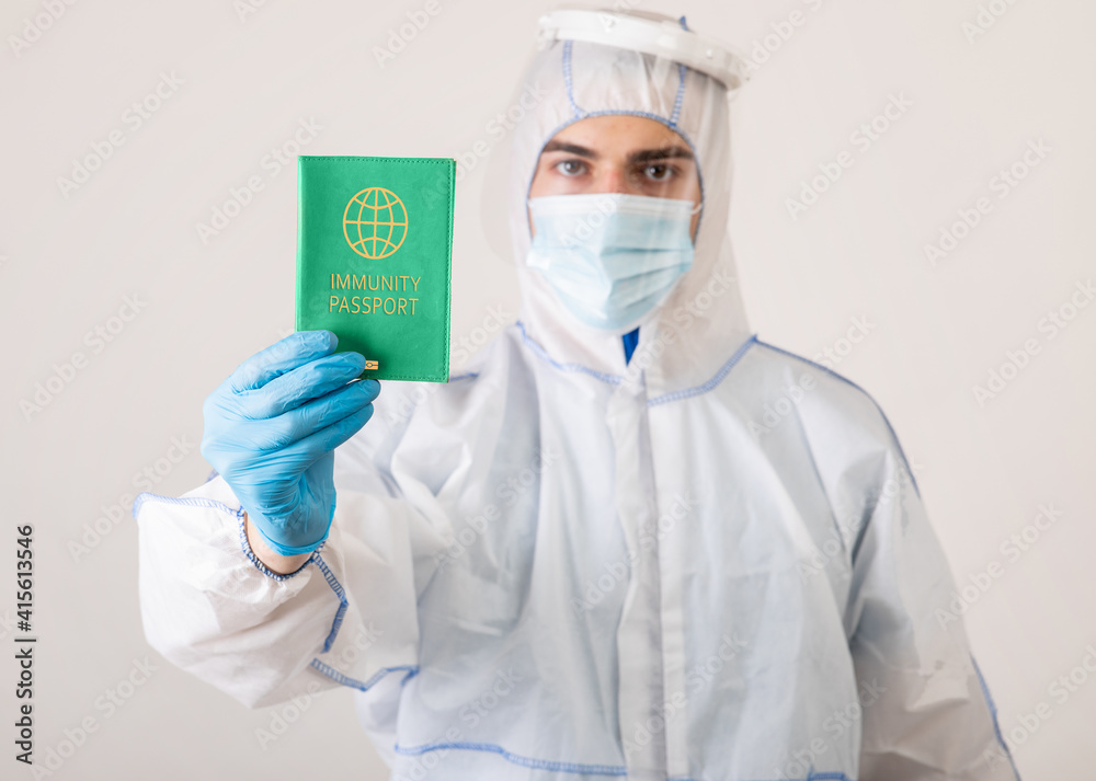Medical specialist in protective biohazard costume showing coronavirus immunity passport on light studio background Medical specialist in protective biohazard costume showing coronavirus immunity passport on light studio background