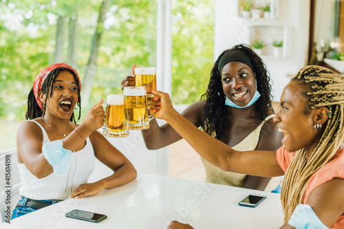 Canvas Print Three young African American girlfriends sitting at brewery (pub) with beer glasses taking off surgical masks