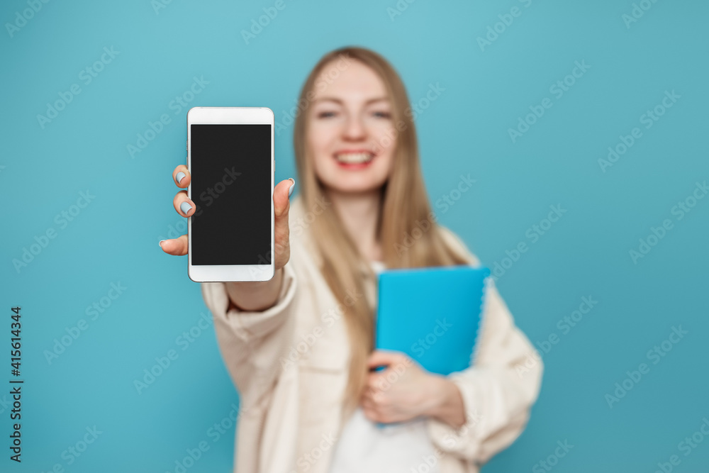 Happy student girl shows a blank screen of a mobile phone at the camera and smiles, girl in blur. Isolated on a blue background in the studio. Mockup