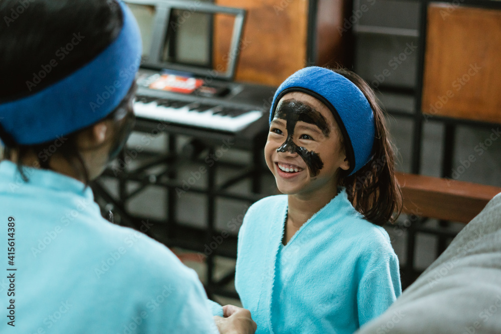 a happy little girl with a black clay mask on half of her face while sitting on the sofa a happy little girl with a black clay mask on half of her face while sitting on the sofa