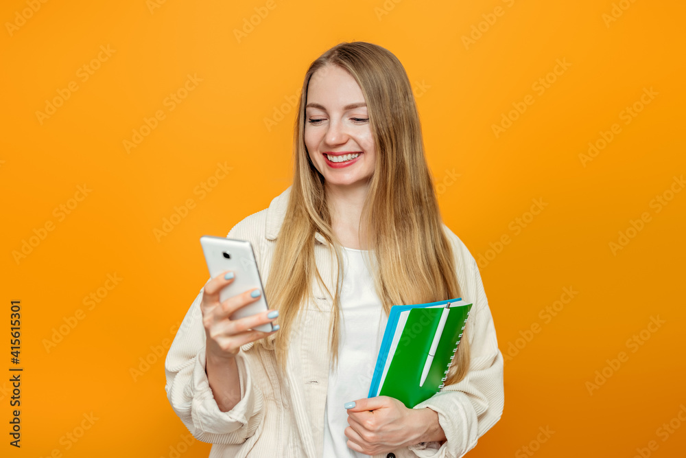 Caucasian blonde student girl looking at the phone screen and smiling isolated over orange studio background. Online education Caucasian blonde student girl looking at the phone screen and smiling isolated over orange studio background. Online education