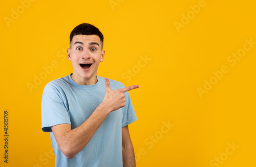 Excited young guy opening mouth in shock and pointing aside at blank space on orange studio background, banner design