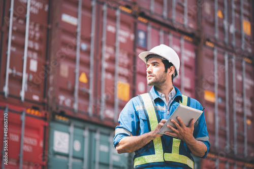 Engineer or foreman holding tablet and wears PPE looking at left side to checking inventory or job details with cargo container background. Engineering site and working with technology concept.