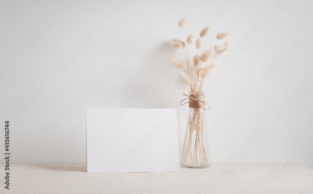Blank white greeting card mock up.decoration with dried Lagurus ovatus flowers composition in modern glass vase  on  beige table and cement wall background