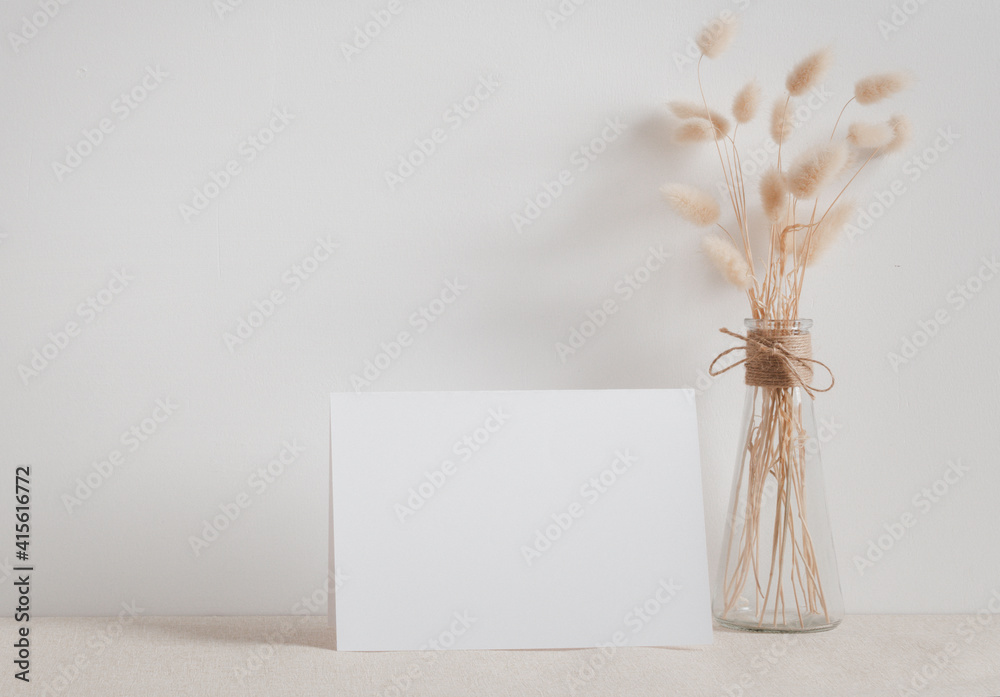 Blank white greeting card mock up.decoration with dried Lagurus ovatus flowers composition in modern glass vase  on  beige table and cement wall background