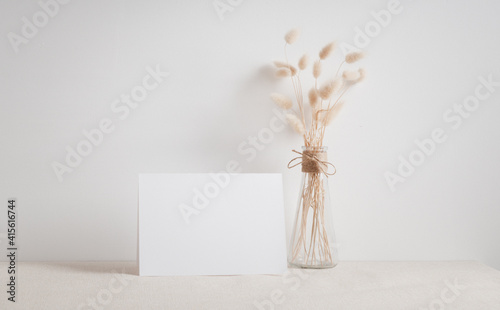 Blank white greeting card mock up.decoration with dried Lagurus ovatus flowers composition in modern glass vase  on  beige table and cement wall background