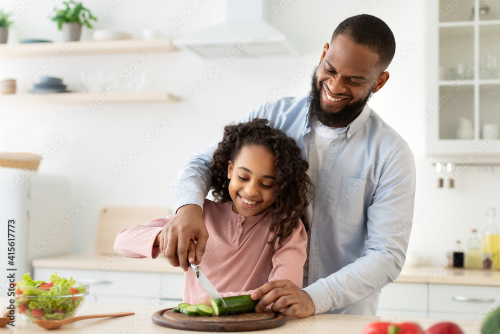 Cheerful african father teaching daughter how to cut veggies