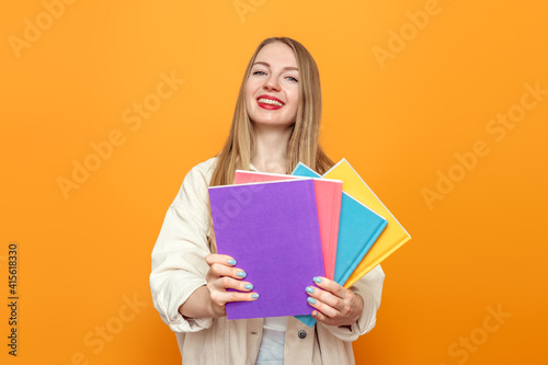 Caucasian blonde student girl in beige jacket holds four books in multi-colored covers smiling isolated over orange background in studio. English language school, education concept