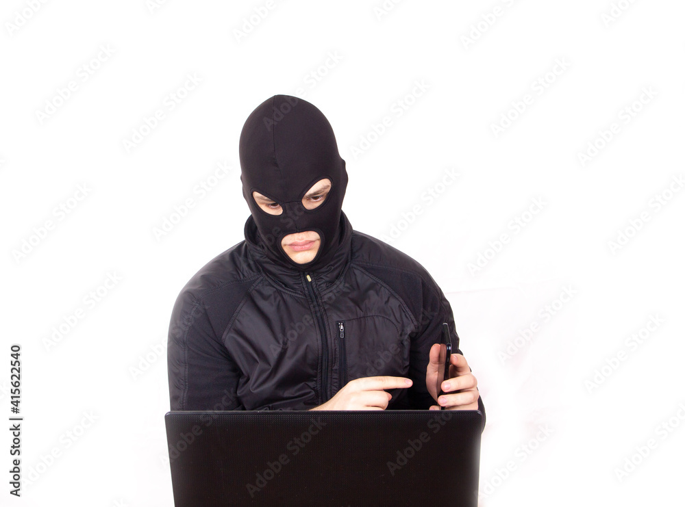 man in black mask with phone and laptop engaged in internet fraud man in black mask with phone and laptop engaged in internet fraud